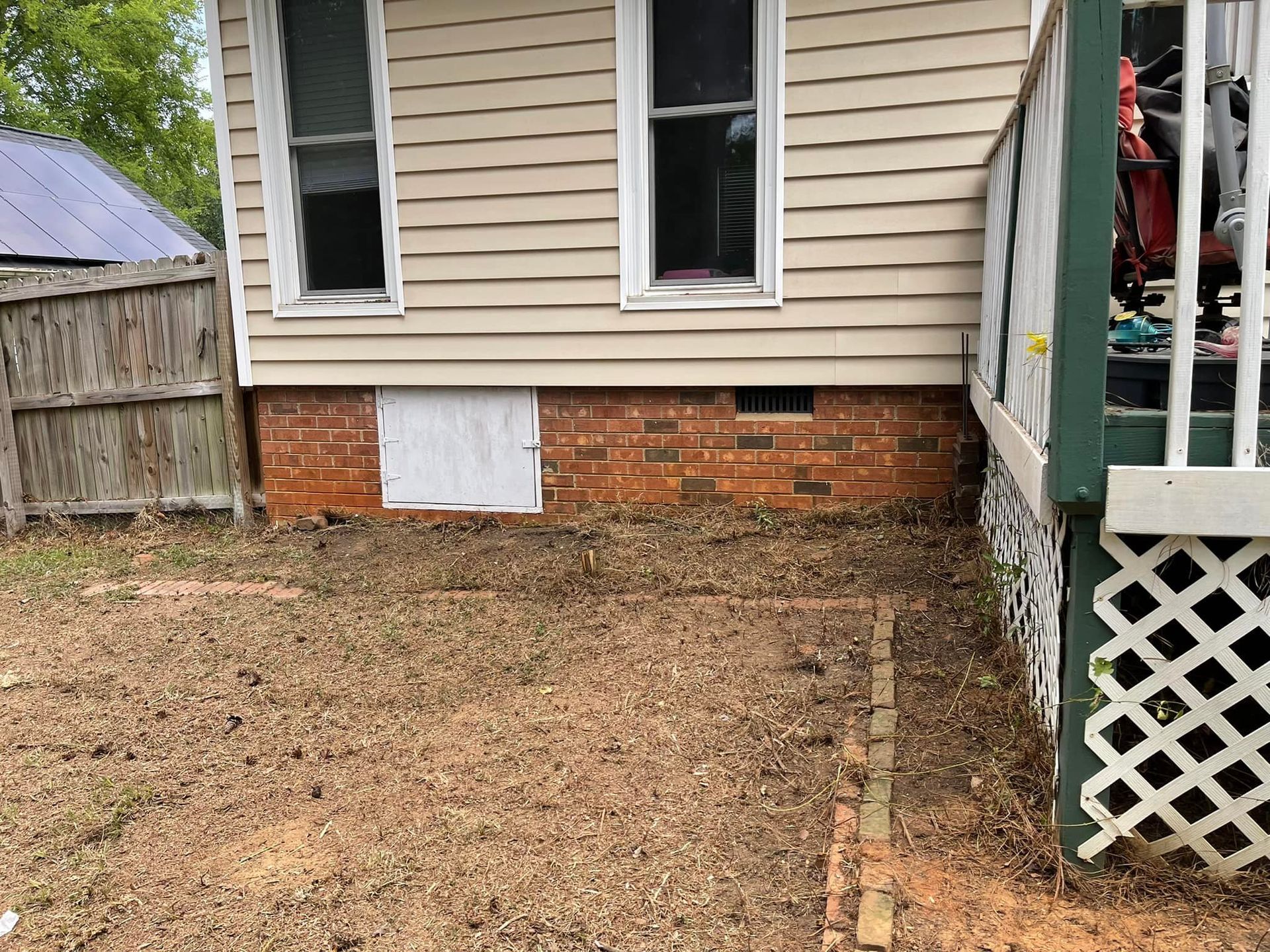 Exterior view of a beige house with a brick foundation, crawlspace door, and a partially visible green wooden deck.