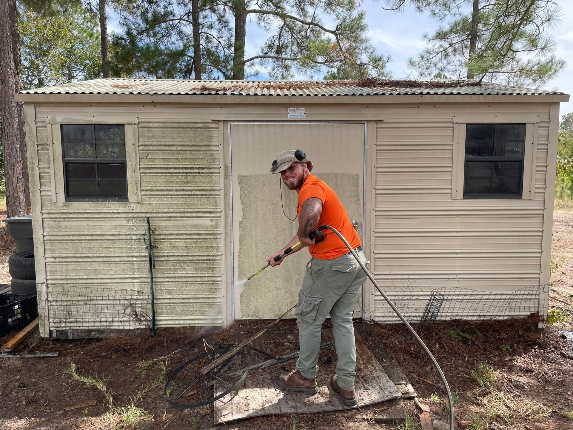 A person in an orange shirt uses a pressure washer to clean the exterior of a weathered, light-colored storage shed.