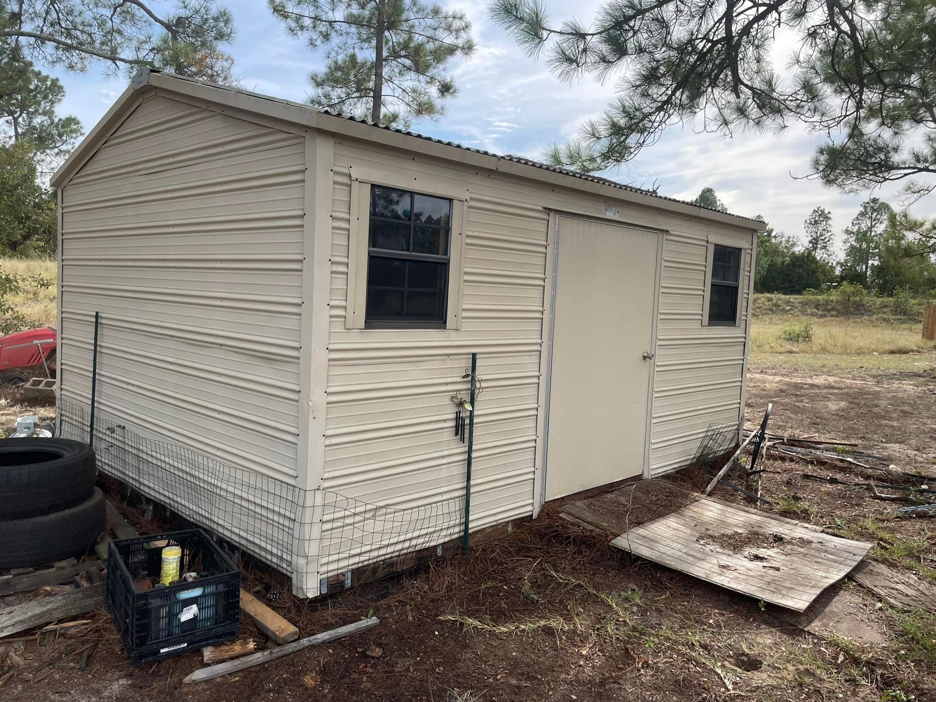 A beige metal storage shed with two small windows and a single door, situated on a dirt lot near trees.
