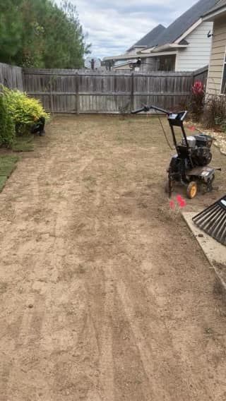 A backyard under renovation with bare, raked soil, a rototiller, and a rake on a concrete patio near a wooden fence.