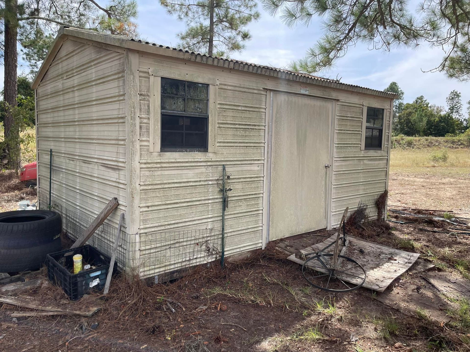 A weathered, beige metal storage shed with two windows and a central door, situated in a grassy, wooded outdoor area.