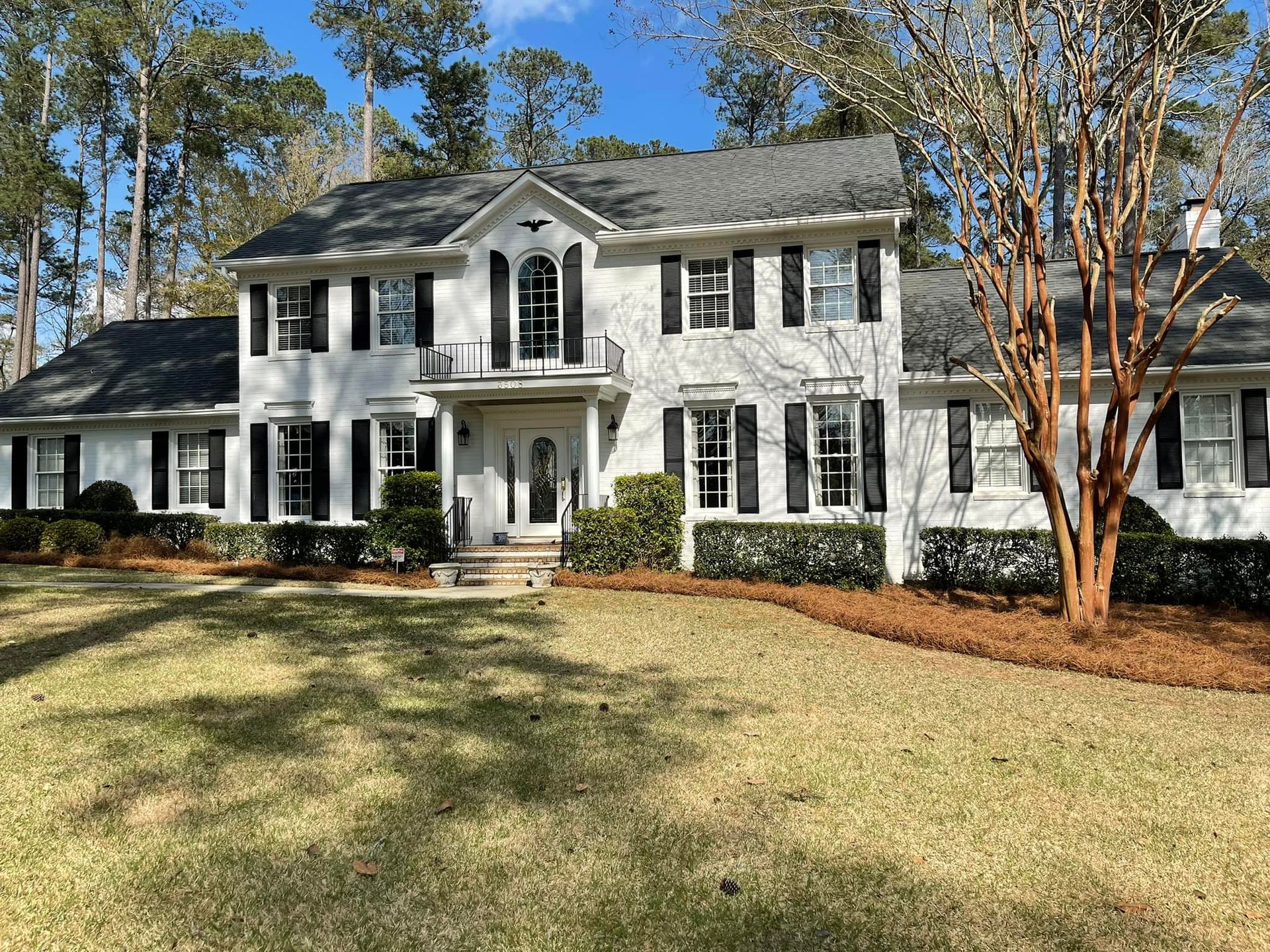 Two-story, white brick house with black shutters and a gray shingled roof, surrounded by trees and a brown lawn.