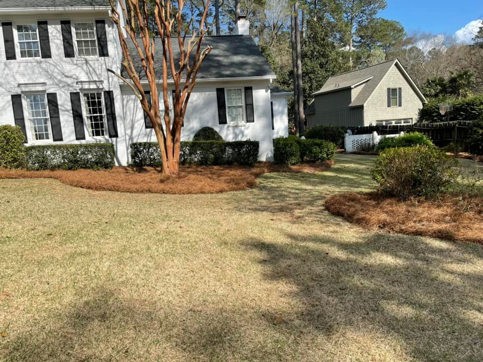 A white two-story house with black shutters, a crepe myrtle tree in the front yard, and a smaller house visible behind it.