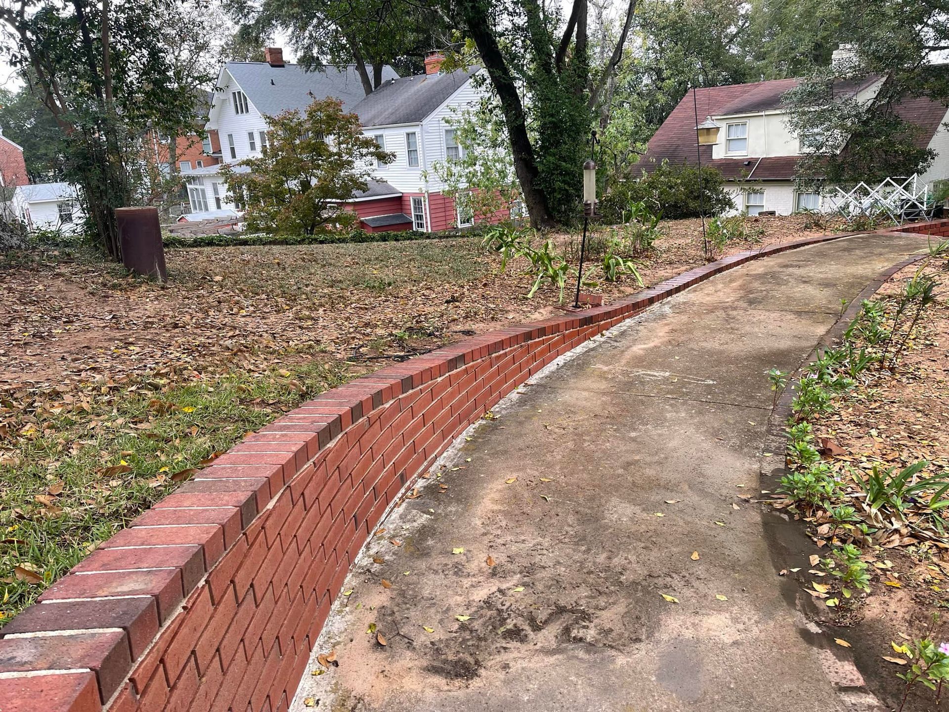 A curved, red brick retaining wall borders a dirt walkway in front of houses on a sunny, leaf-strewn residential lot.