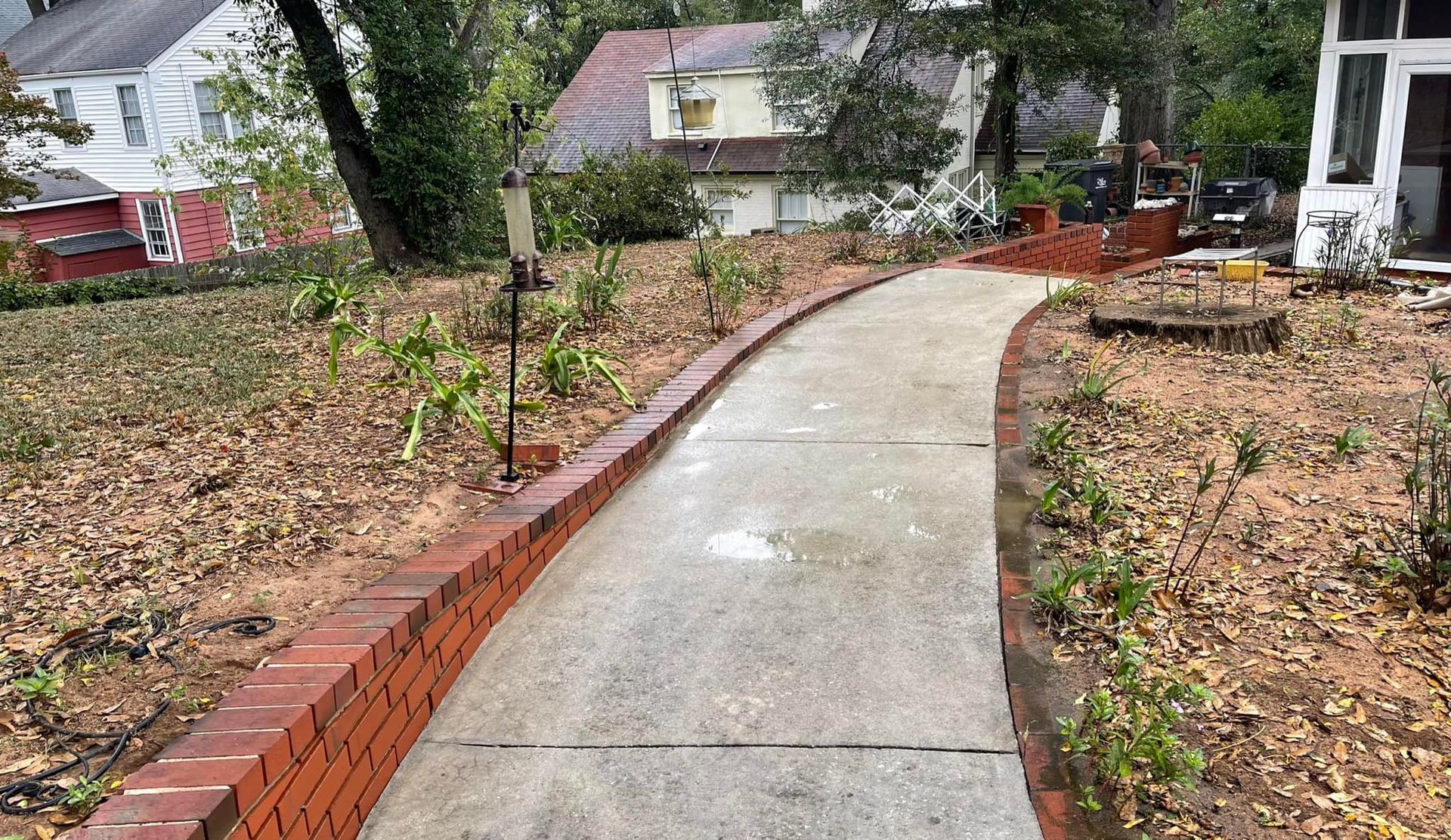 A concrete sidewalk winds through a yard with a brick retaining wall on the left, set against a backdrop of houses.