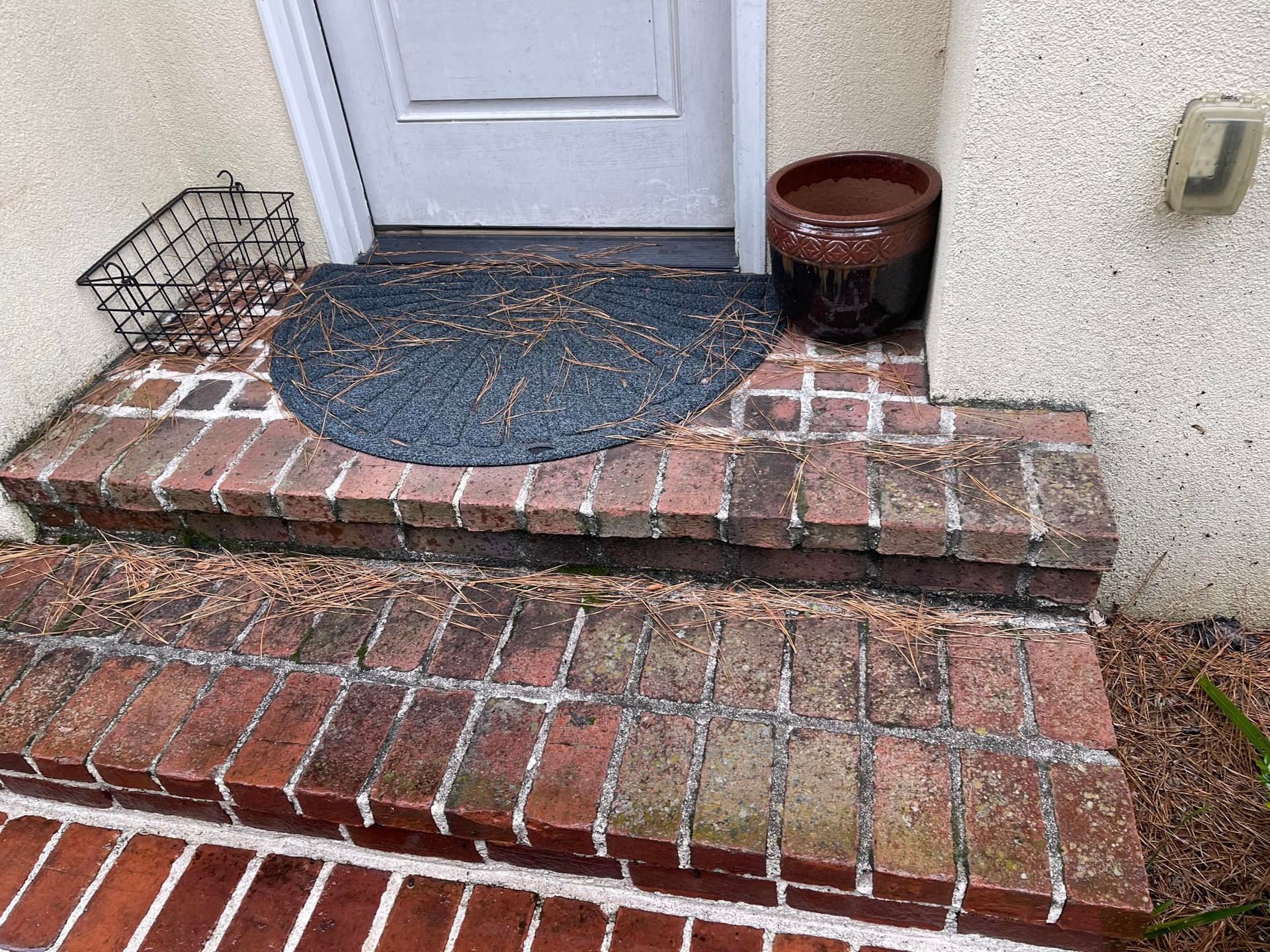 Brick steps leading to a white door with a semi-circular mat, a wire basket, and a brown planter.