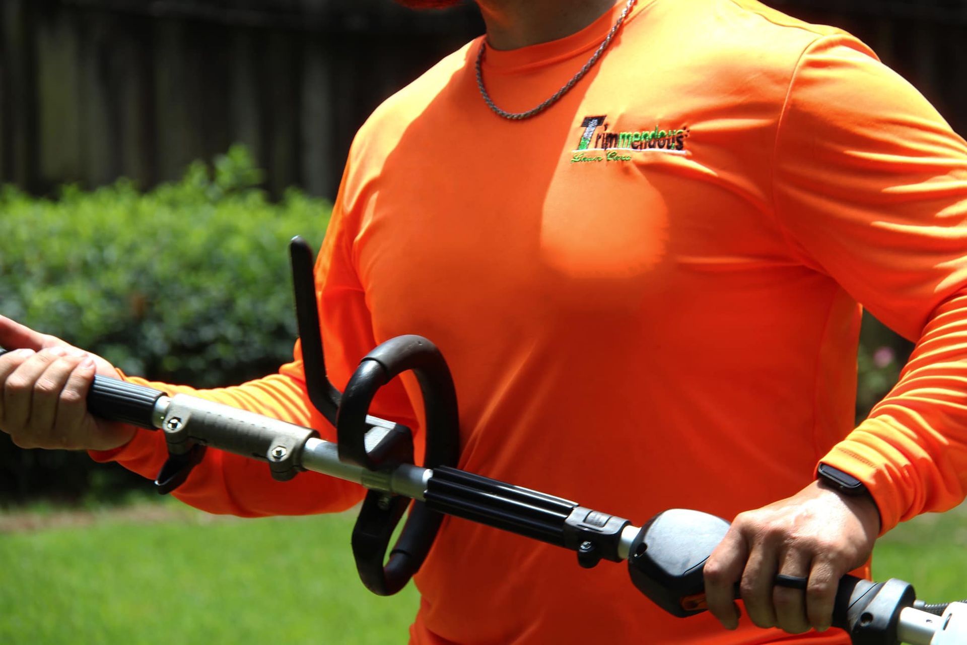 A person in a bright orange long-sleeved shirt holds a weed trimmer outdoors, set against a green, grassy background.