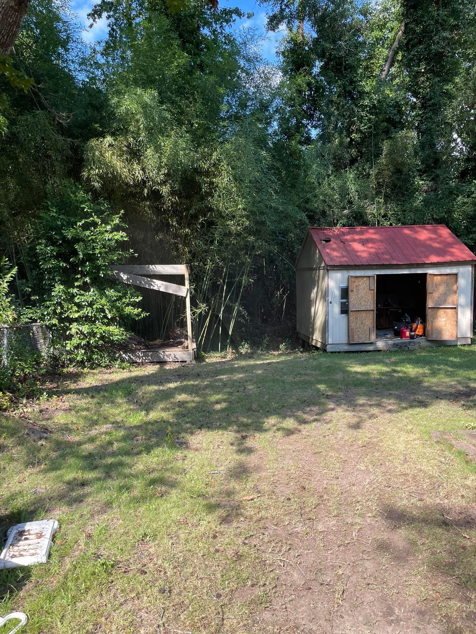A small white shed with a red roof stands in a grassy yard beside a dense line of trees, with a partially obscured structure.