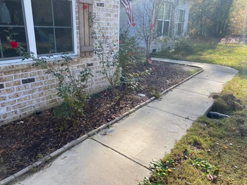 A concrete walkway leads past a brick house exterior with a garden bed featuring rose bushes and mulch.