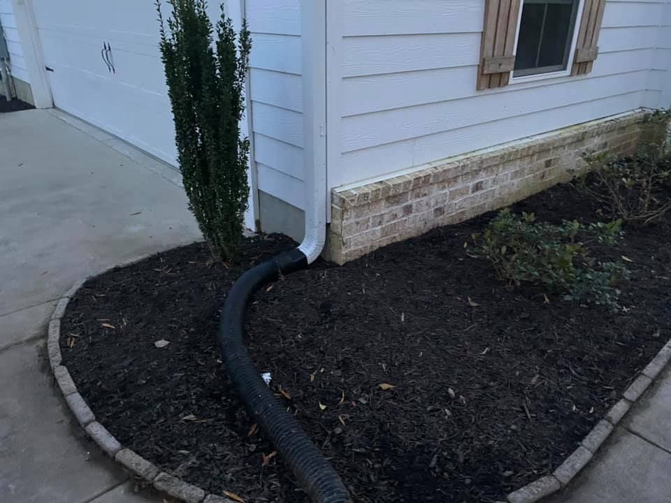 A black flexible downspout extension directs water from a white house corner into a landscaped mulch bed.