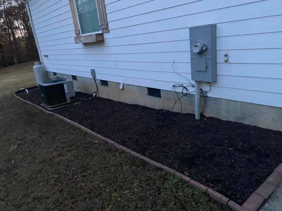 A house exterior with white siding, an electrical meter, a black HVAC unit, and a mulched garden bed with brick edging.