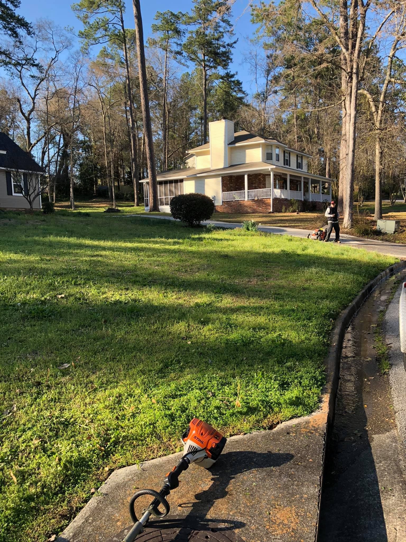 A string trimmer rests on a concrete curb in front of a large, two-story house with a sprawling lawn and trees.