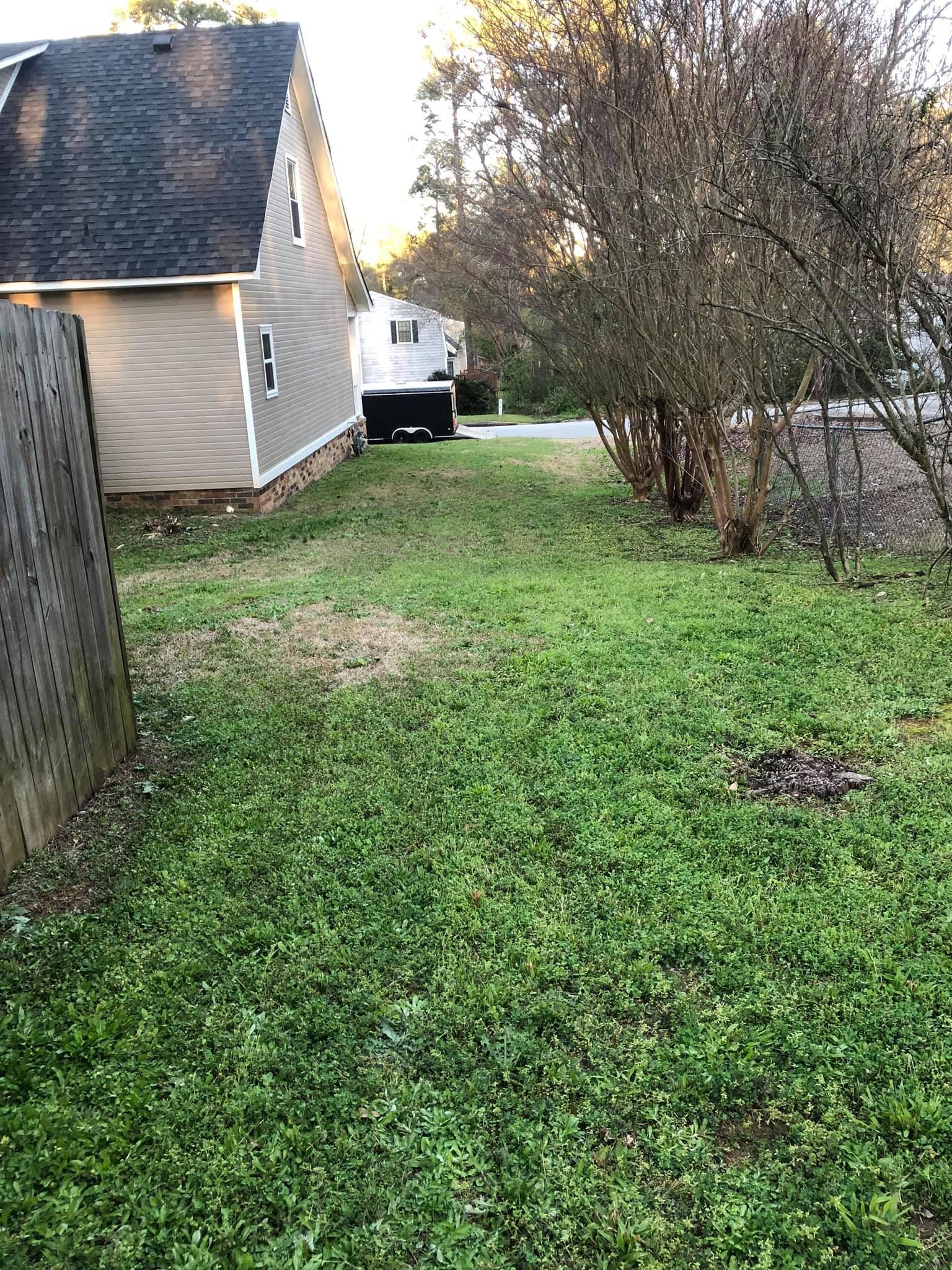 A grassy side yard between a tan house with a dark roof and a fence, with trees and another building in the distance.
