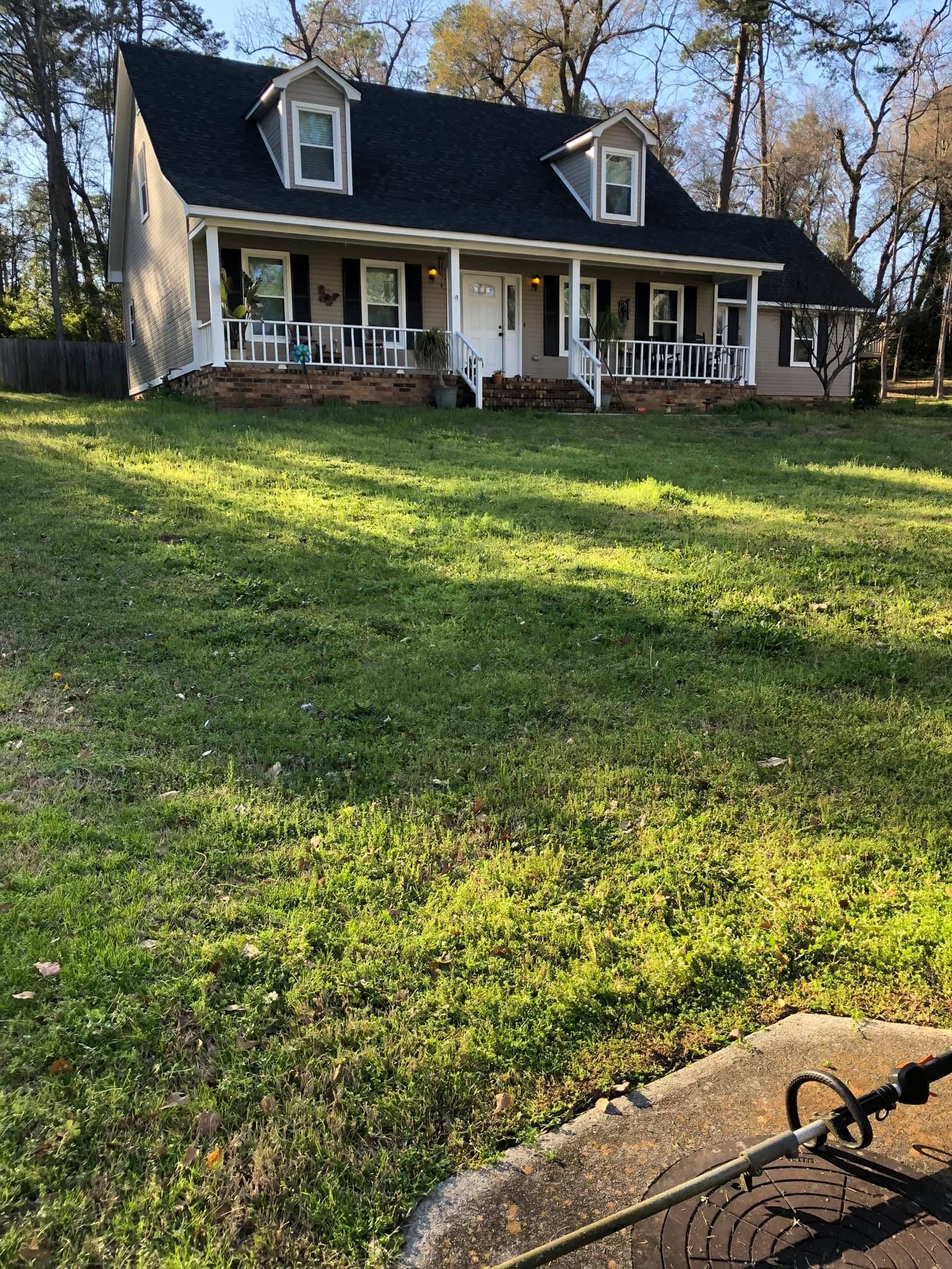 A two-story house with a white porch, dark roof, and dormer windows, situated behind a large, grassy front yard.