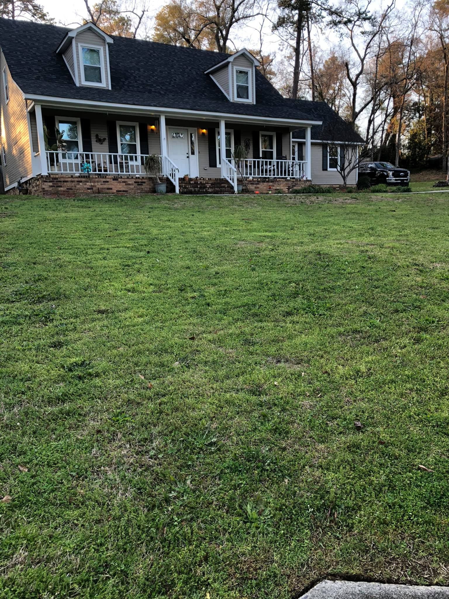 A two-story farmhouse with a covered front porch and dormer windows, set behind a large, green grassy yard.