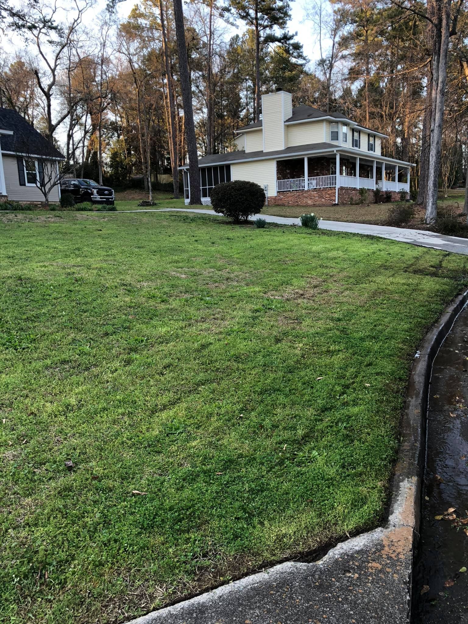 A grassy lawn in front of a two-story beige house with a wrap-around porch, trees in the background, and a sidewalk.