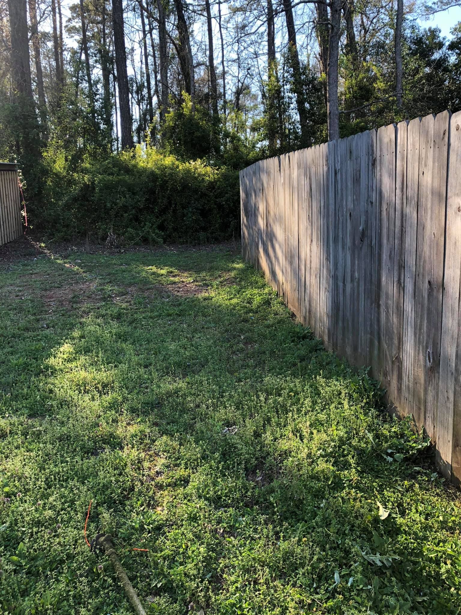 A weathered wooden privacy fence runs along the side of a grassy backyard lined with trees and shrubs.