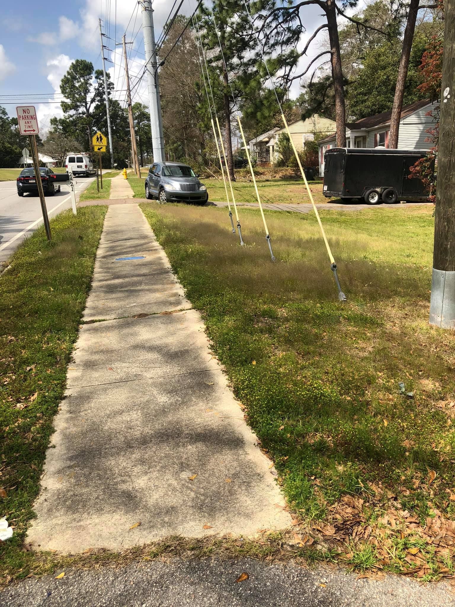 A concrete sidewalk stretches into the distance, flanked by grassy yards with trees, a parked SUV, and a black trailer.