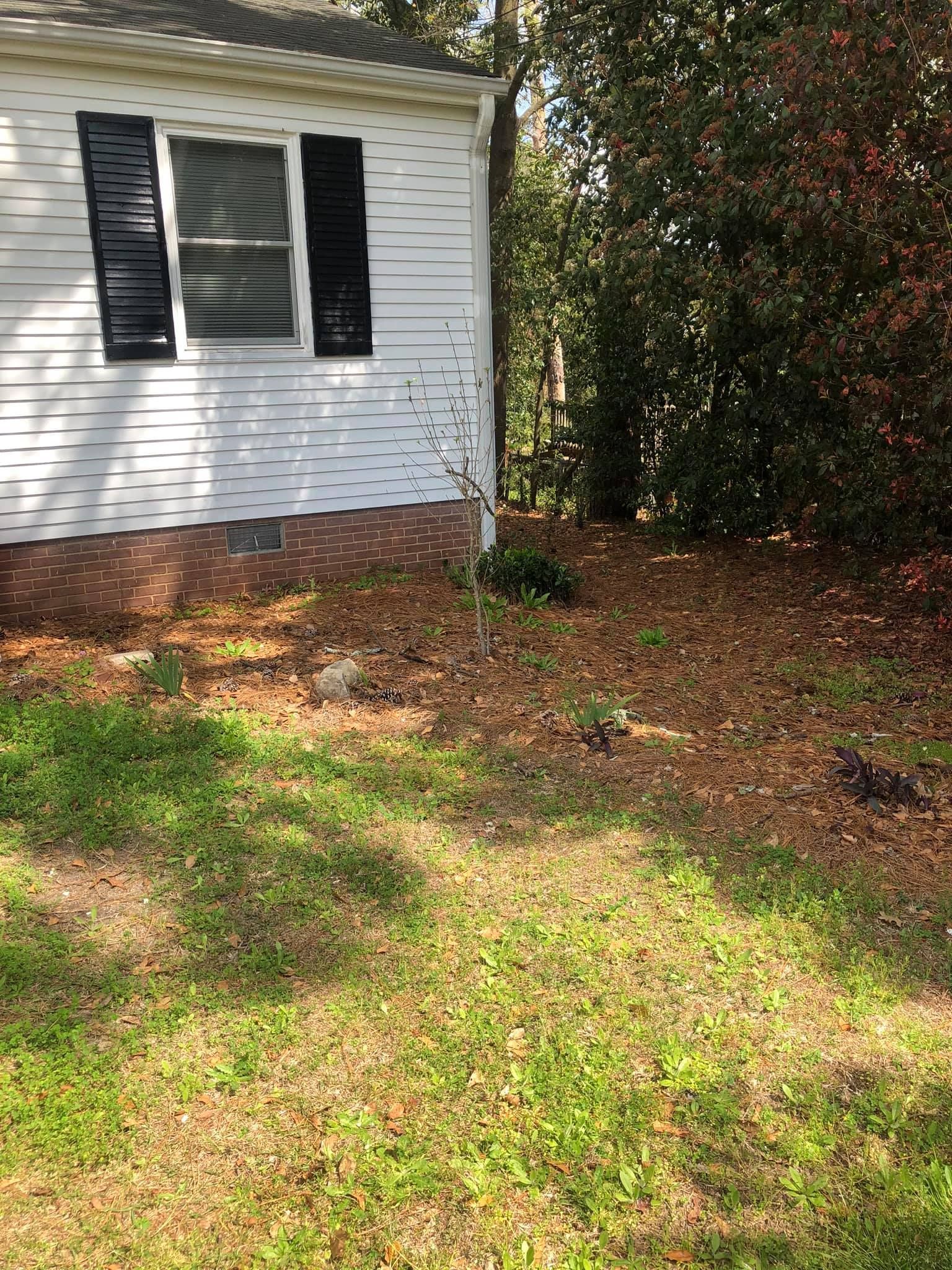 A white house with black shutters and a brick foundation sits next to a wooded area covered in fallen leaves.