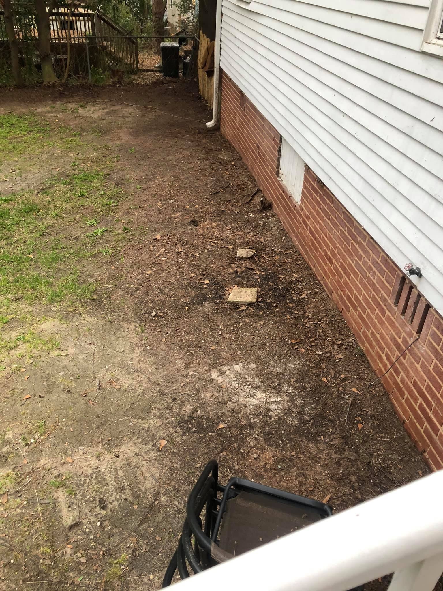 The side of a white-sided house with a brick foundation sits above a dirt yard with scattered leaves and a small chair.
