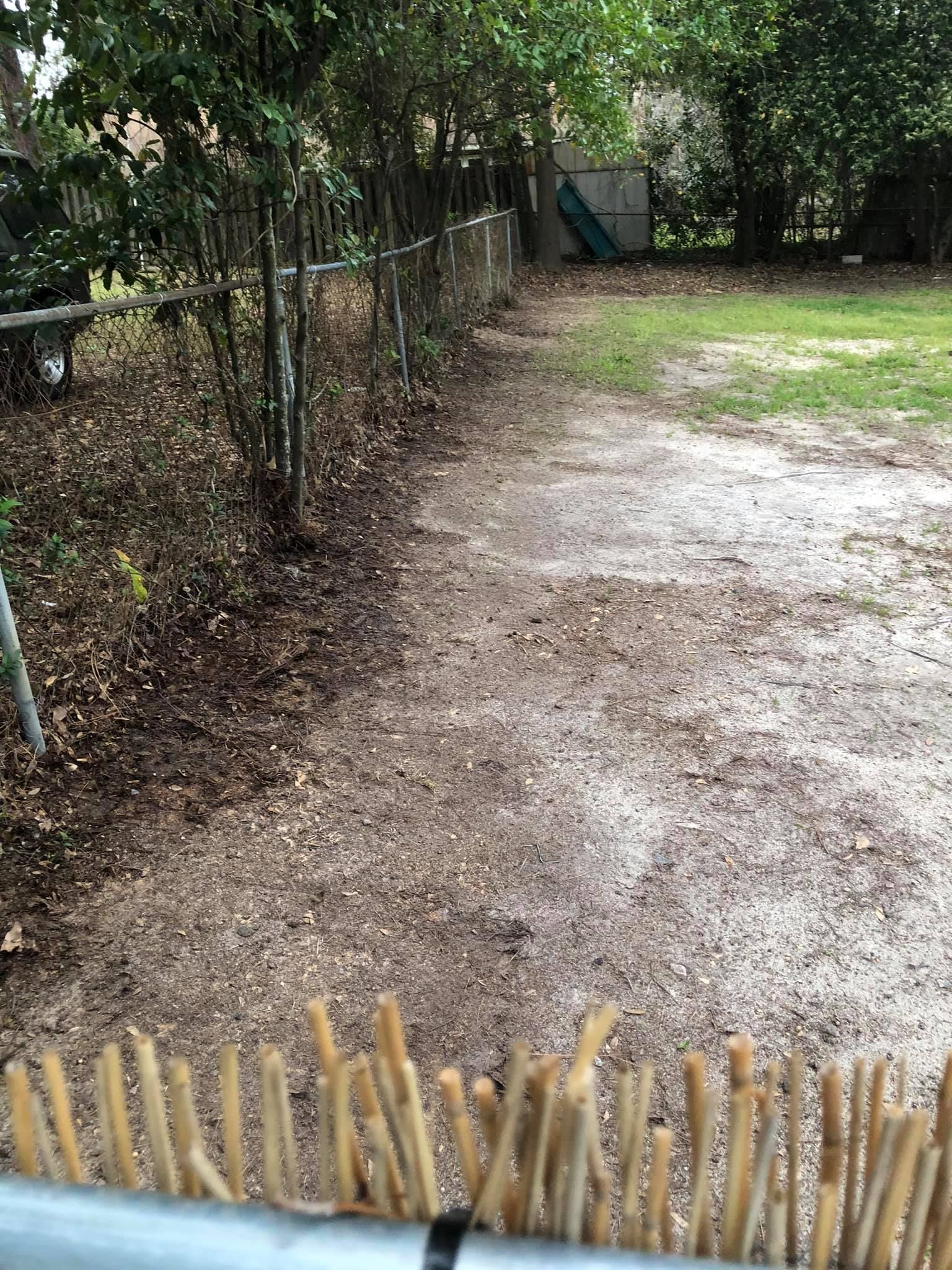 A backyard area showing a dirt and gravel path alongside a chain-link fence, with trees and grass in the background.