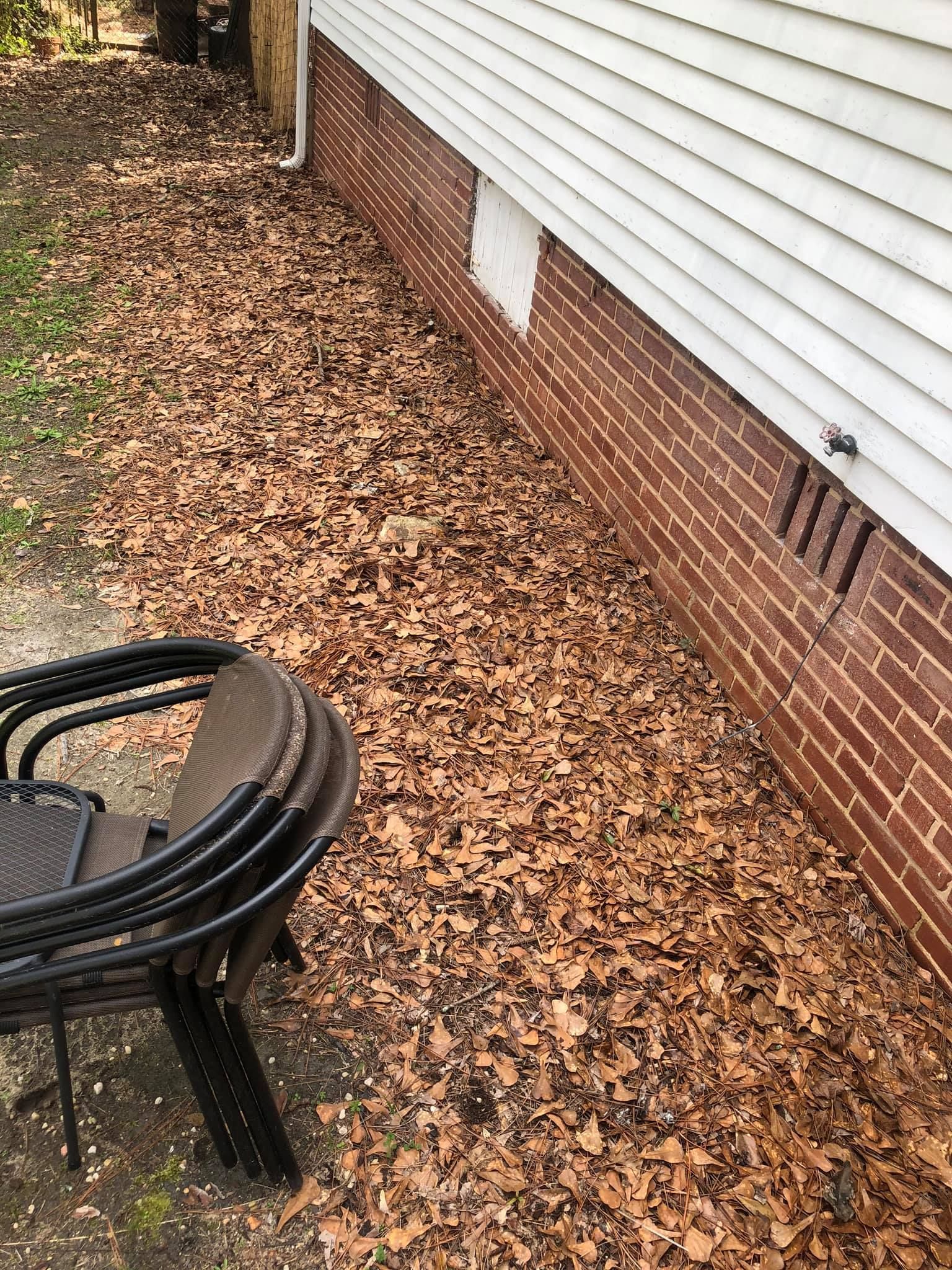 A stack of dark outdoor chairs sits on a concrete patio next to a red brick house foundation covered in fallen brown leaves.