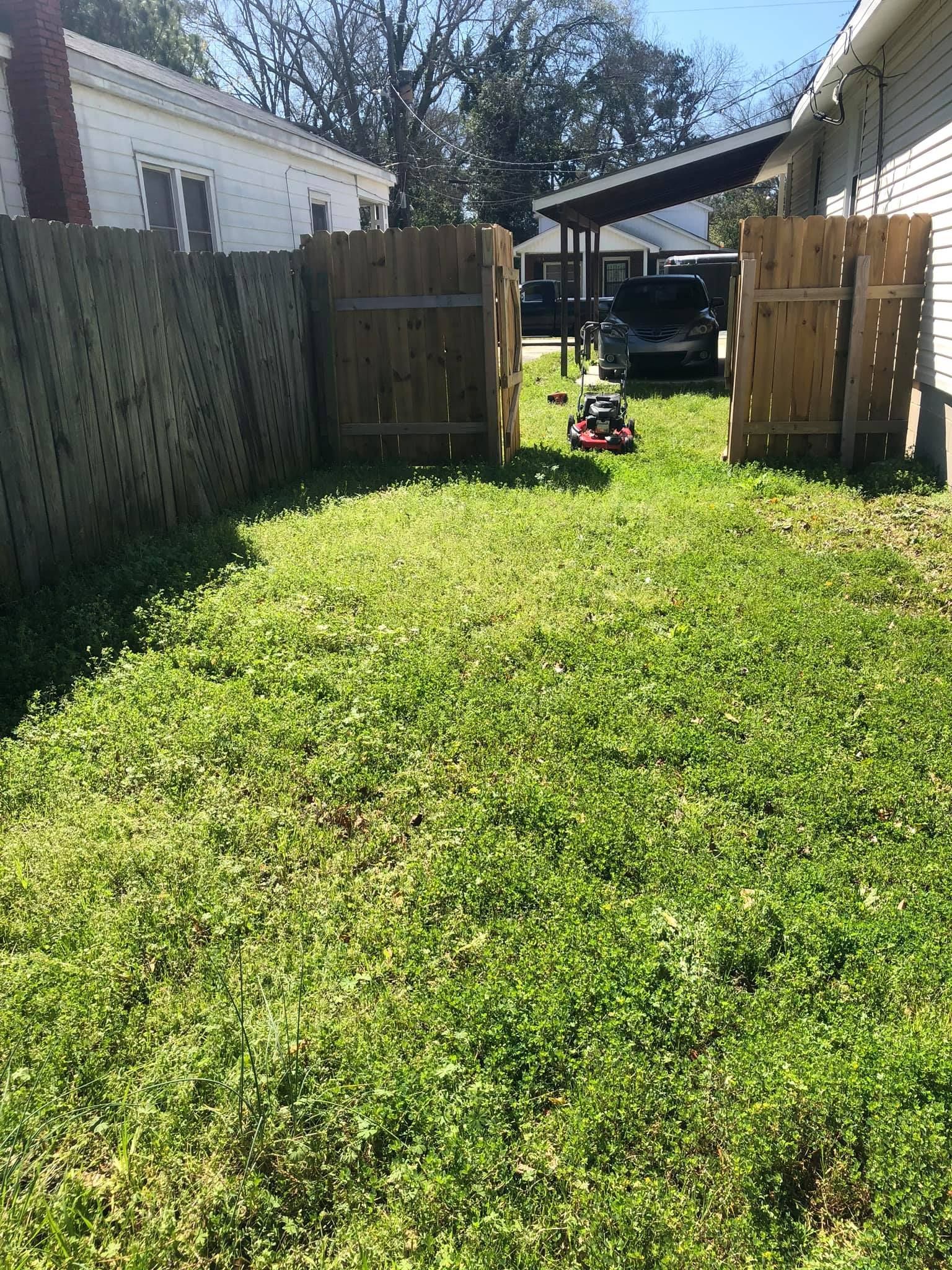 A lawnmower sits on a grassy path between two wooden fences and houses.