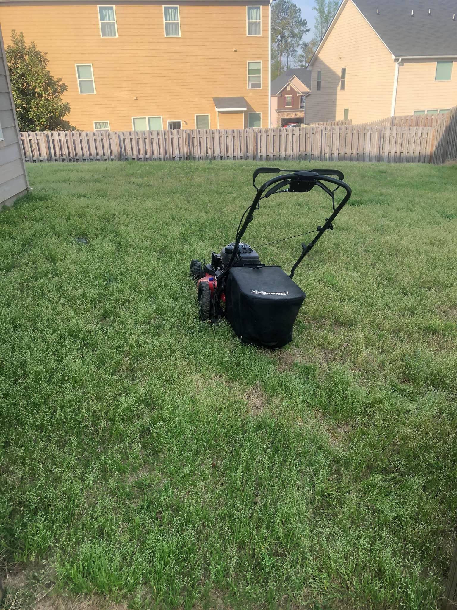 A black lawnmower sits in the middle of a grassy residential backyard with a fence and houses in the background.