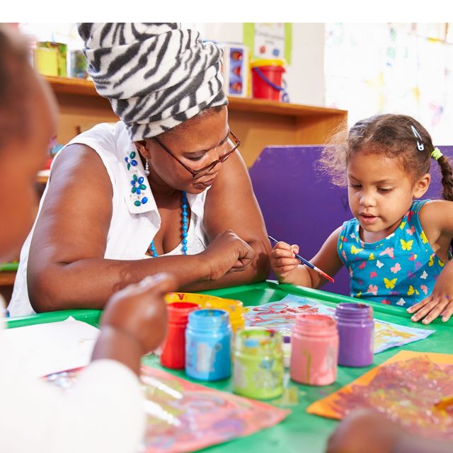 Adult and child painting together at a table with colorful paint jars in a bright classroom