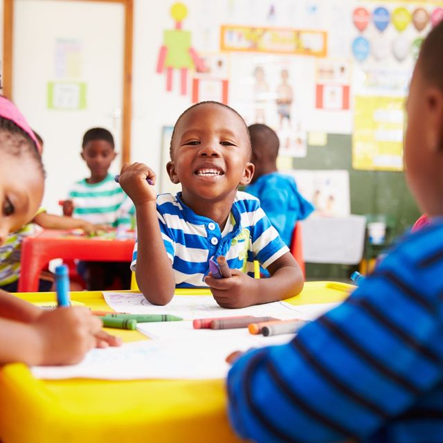 Children smiling and coloring at a bright classroom table with crayons and paper