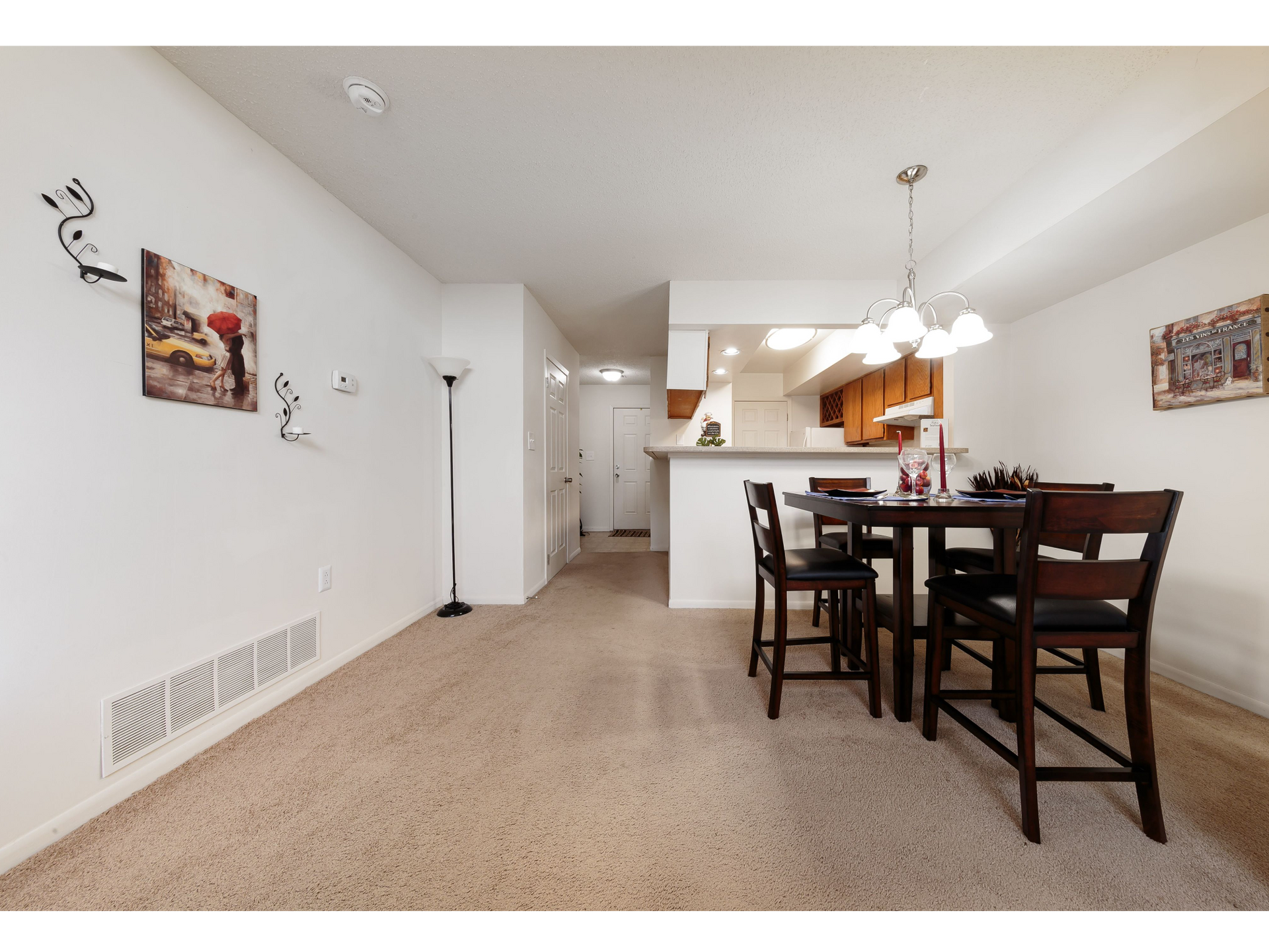 Empty beige dining room and kitchen with wooden table, chairs, and wall art.