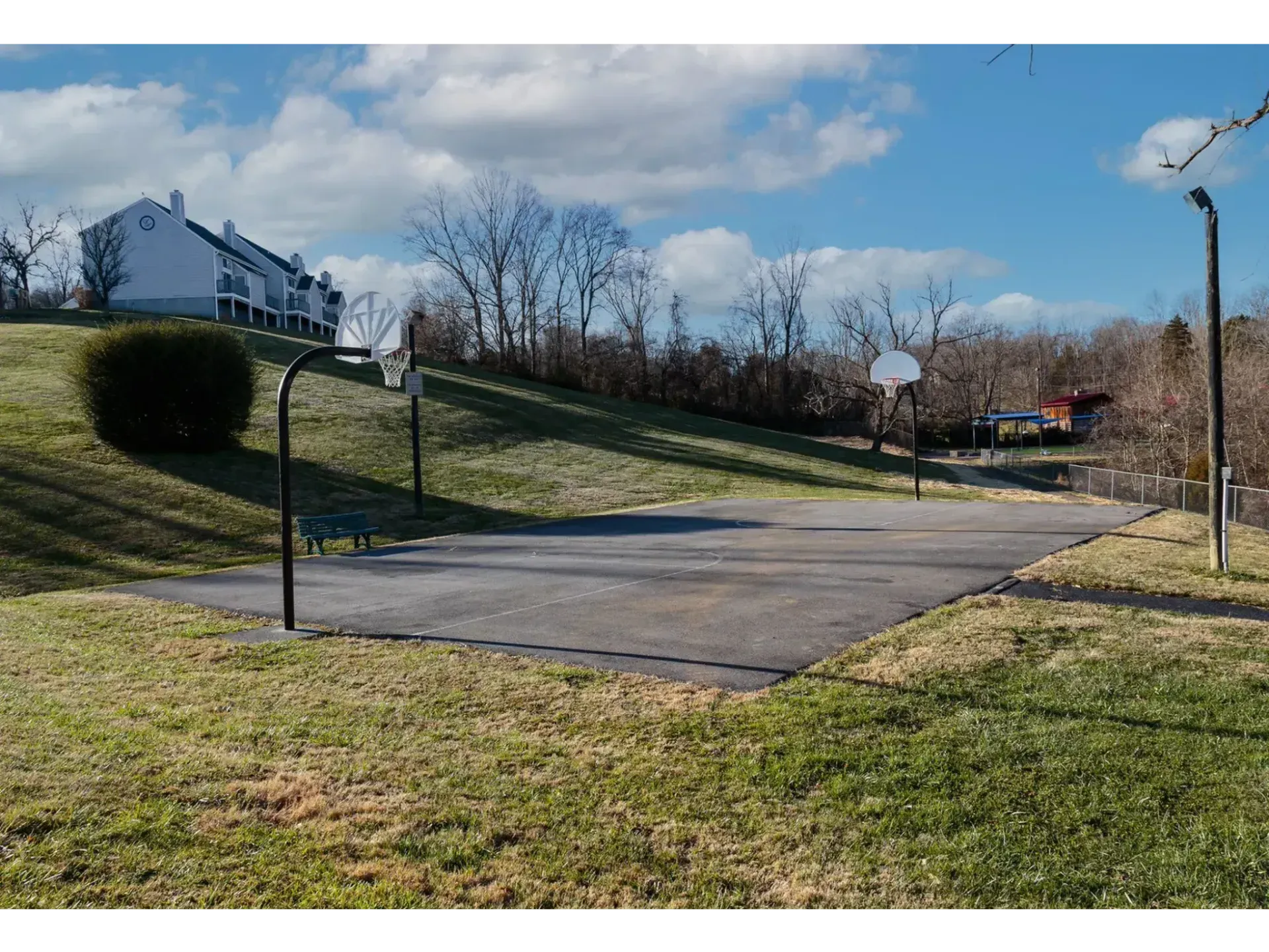 Empty outdoor basketball court beside a grassy hillside with houses and bare trees under a blue sky