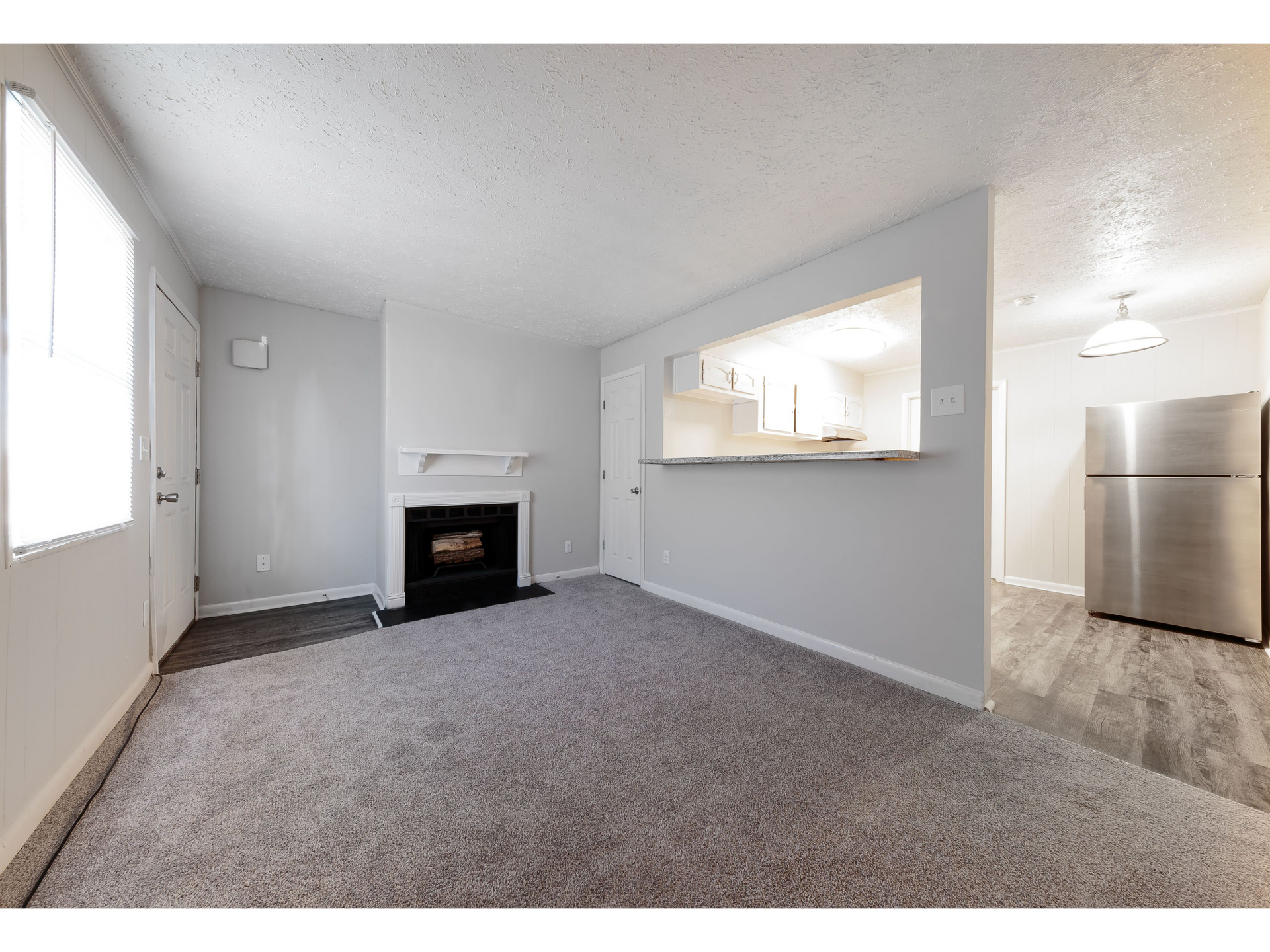 Empty living room with gray carpet, fireplace, pass-through opening, and kitchen area with stainless fridge.