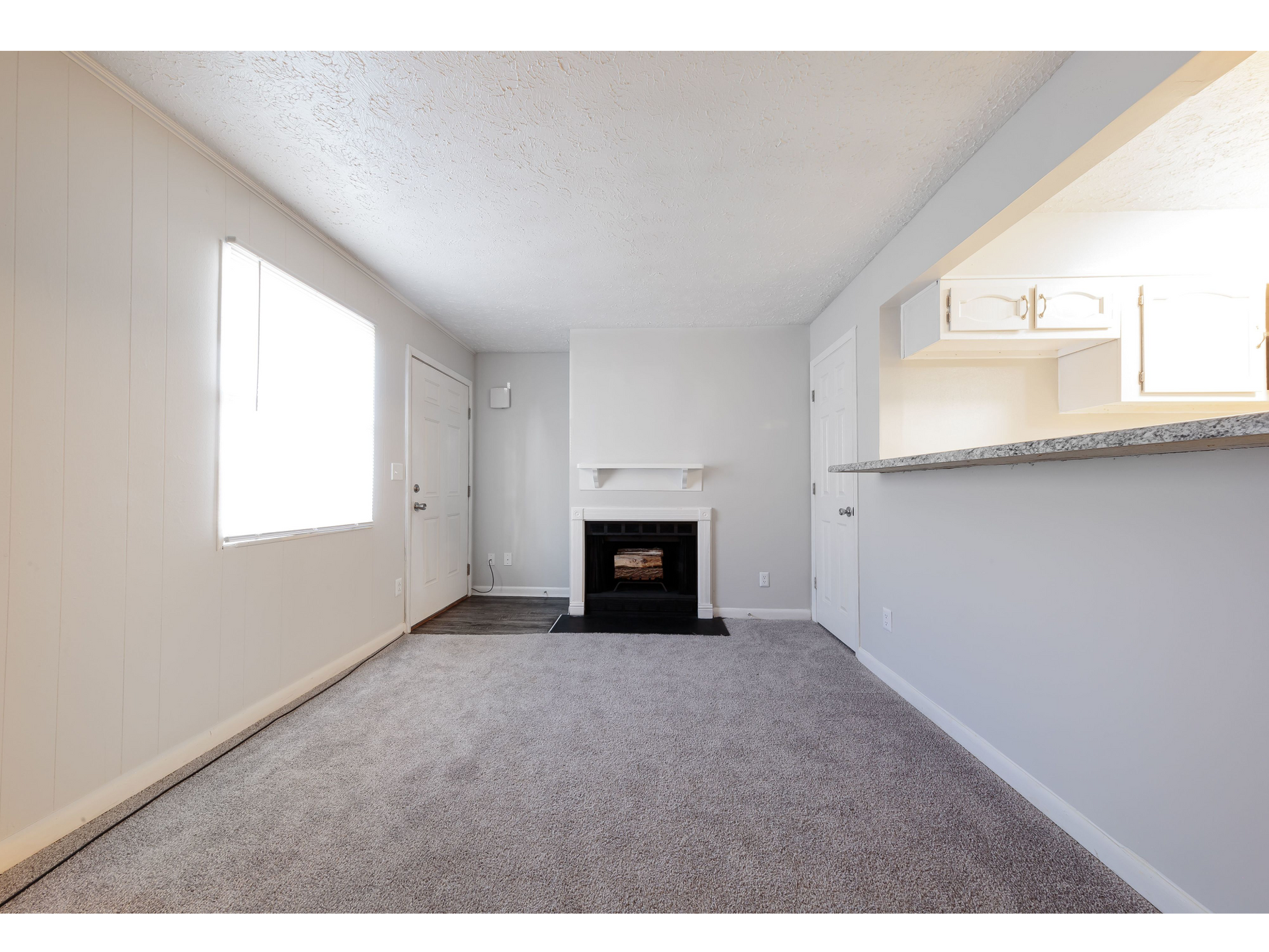 Empty carpeted living room with white walls, a fireplace, and a kitchen pass-through counter