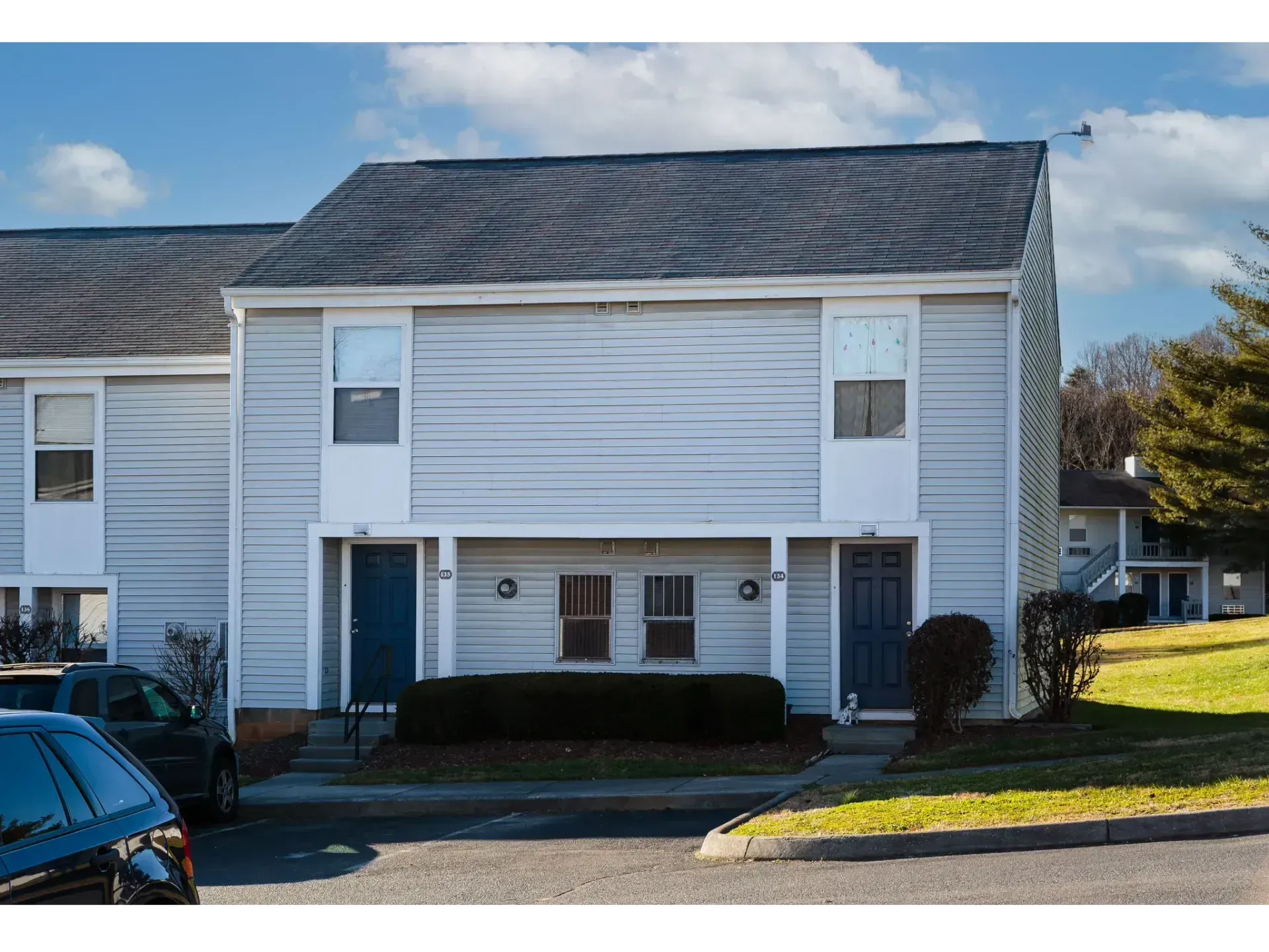 Two-story white townhouse with gray roof, dark blue doors, and shrubs in front under a cloudy sky
