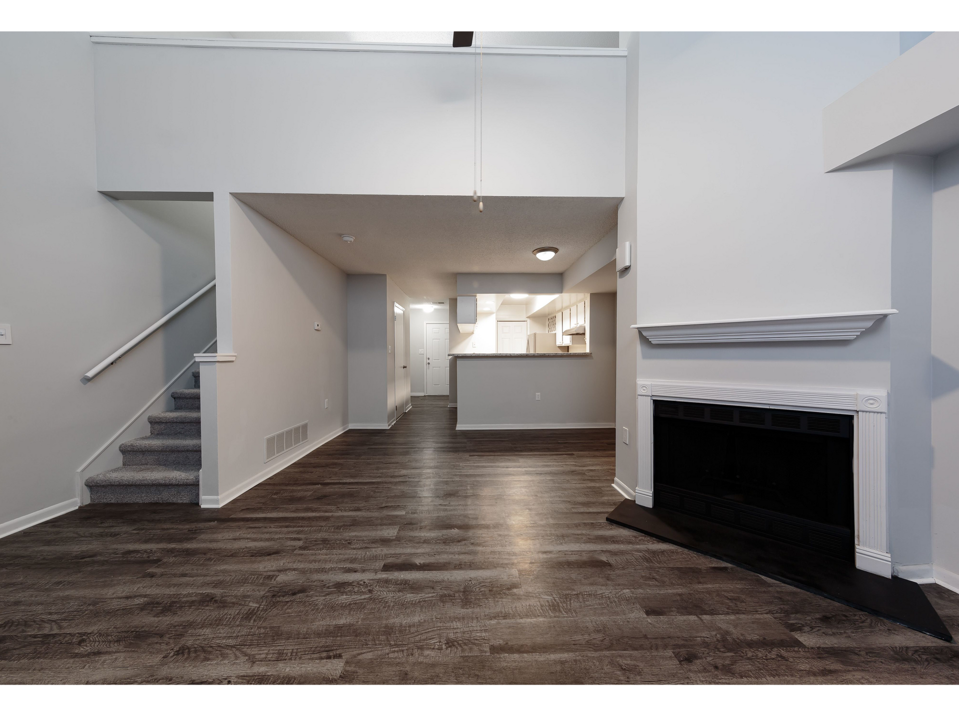 Empty modern living room with dark wood floors, white walls, kitchen pass-through, stairs, and fireplace