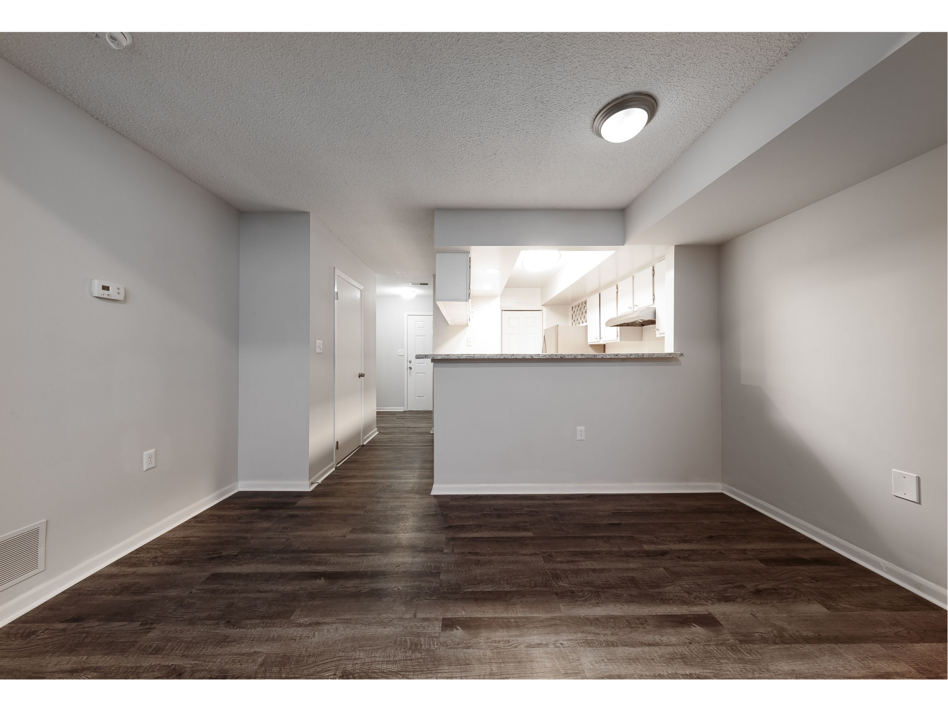 Empty apartment living room with dark wood floors and a small kitchen pass-through, white walls and ceiling lights