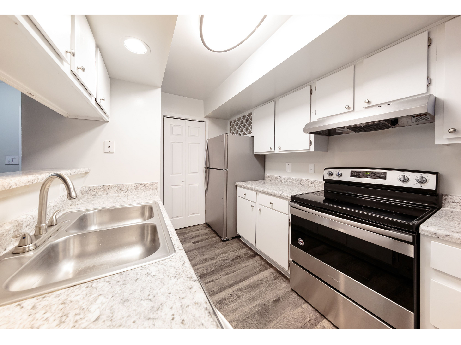 Bright modern galley kitchen with white cabinets, granite counters, stainless sink, fridge, and stove.