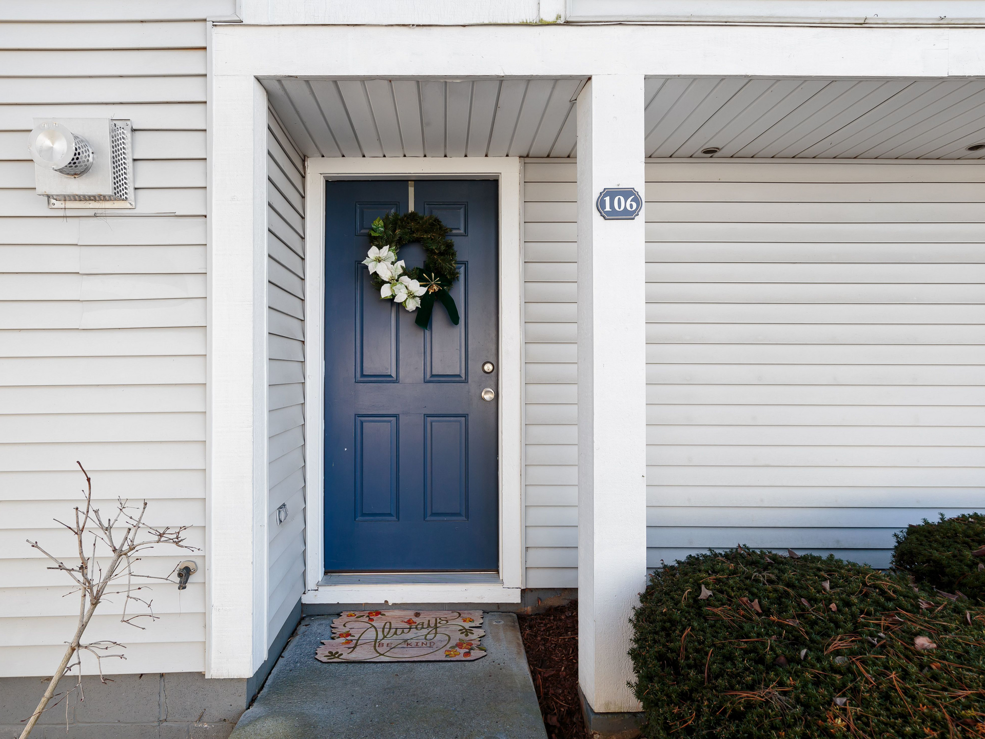 Blue front door with a holiday wreath, framed by white siding and a small welcome mat.