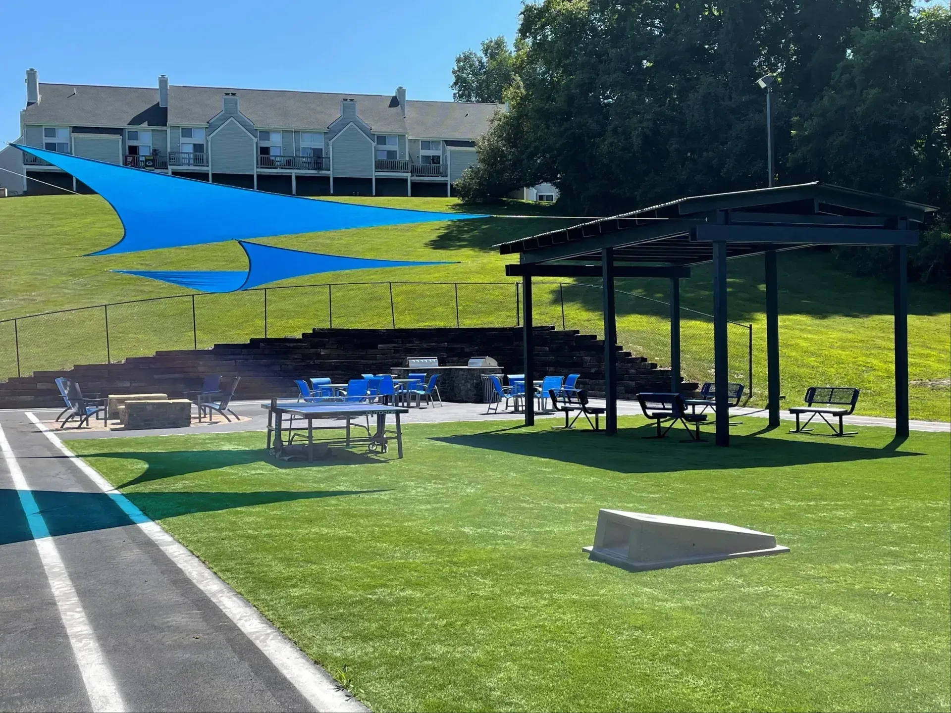 Outdoor picnic area with blue shade sails, tables, pavilion, and grassy hill beside apartment buildings.