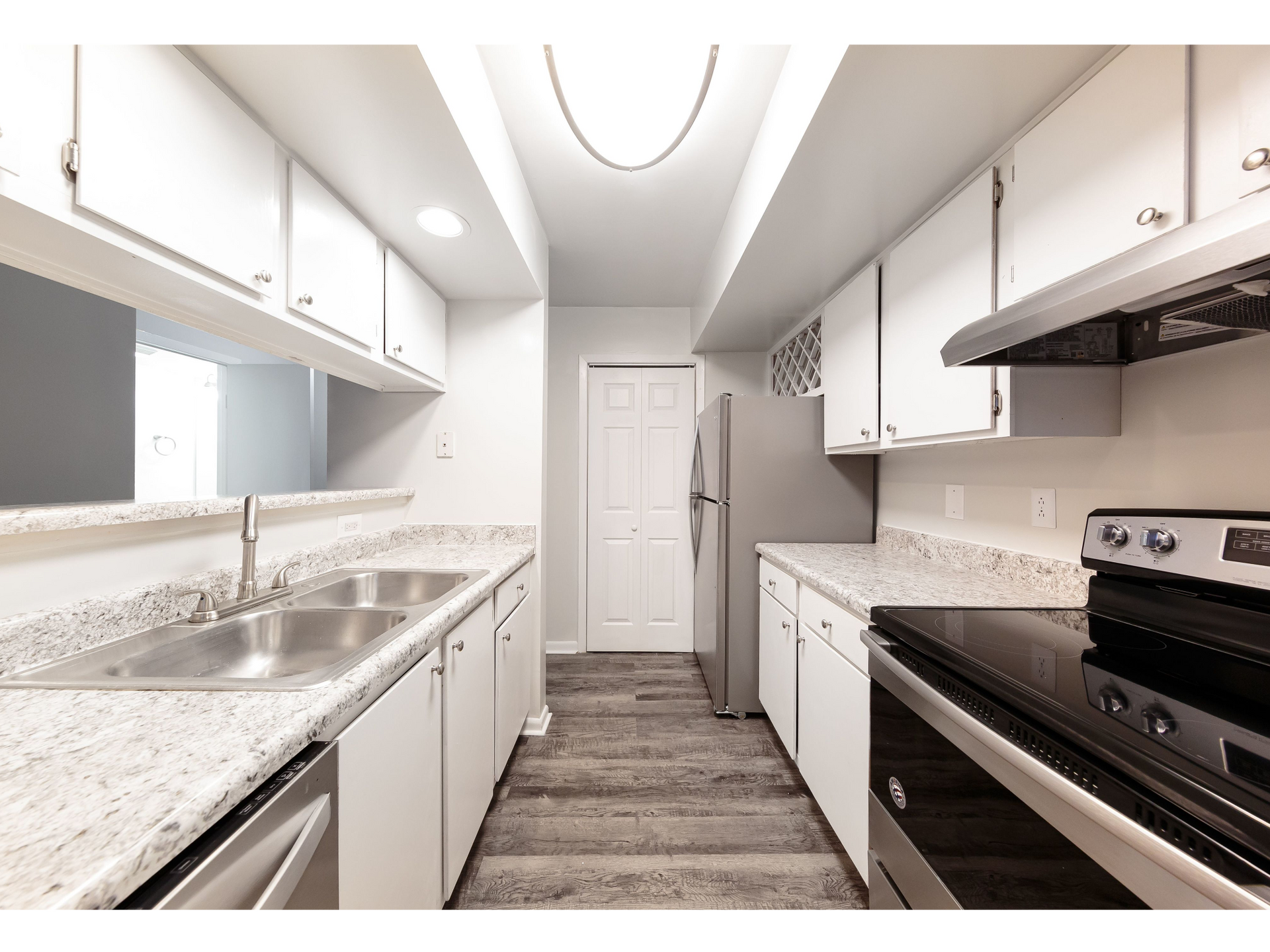 Bright galley kitchen with white cabinets, granite countertops, stainless appliances, and wood-look flooring