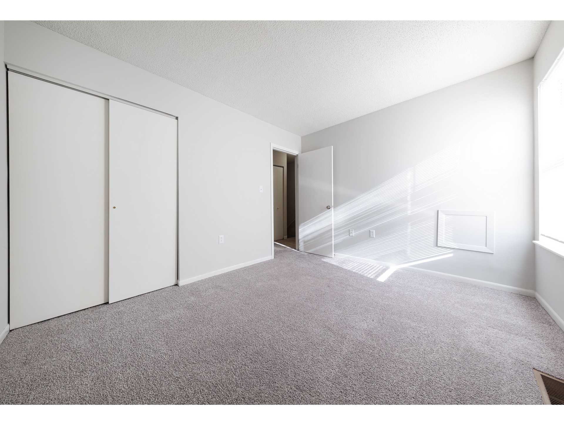 Empty carpeted bedroom with white walls, closet doors, and sunlight streaming through blinds