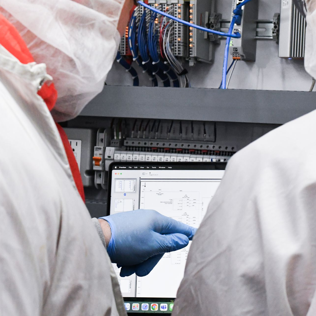 Two people in white protective suits and blue gloves working on a control panel with a laptop in a factory setting.