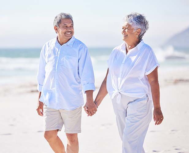 Two people holding hands and walking on a sunny beach, smiling at each other.
