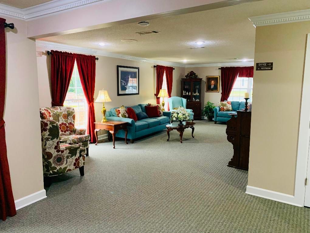 Bright lounge with teal sofas, floral armchairs, red curtains, and framed art in a carpeted room.