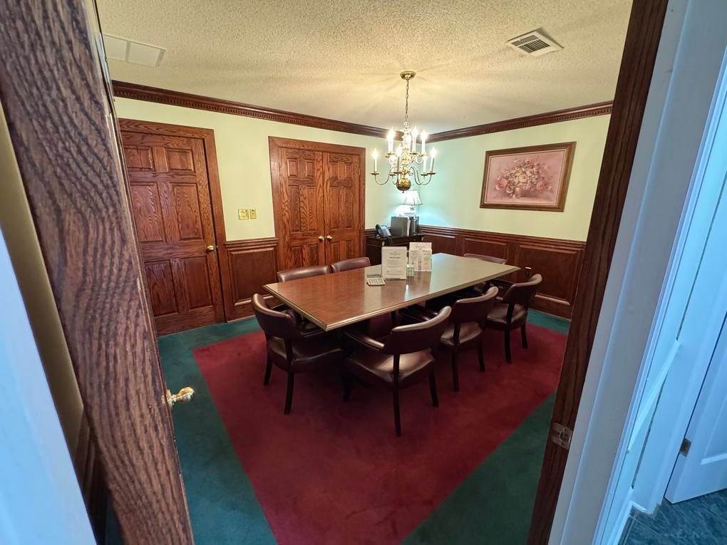 Dining room with a long wooden table, burgundy carpet, and framed wall art under a chandelier.