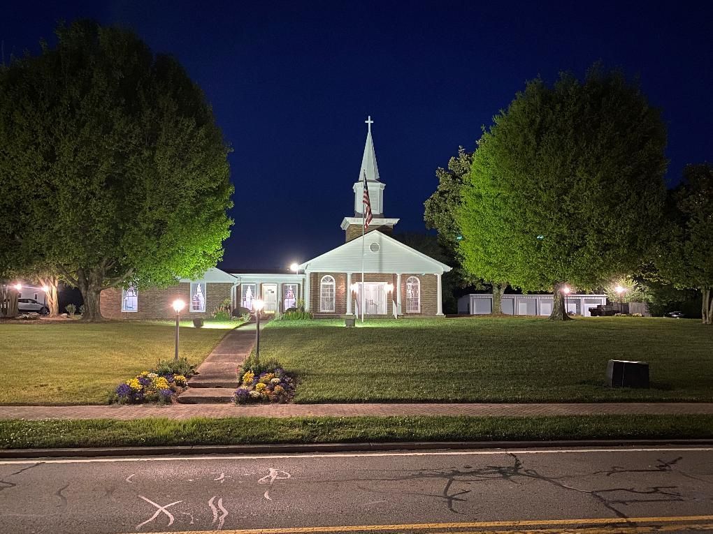 Illuminated white church at night with a steeple, framed by large trees and a front walkway