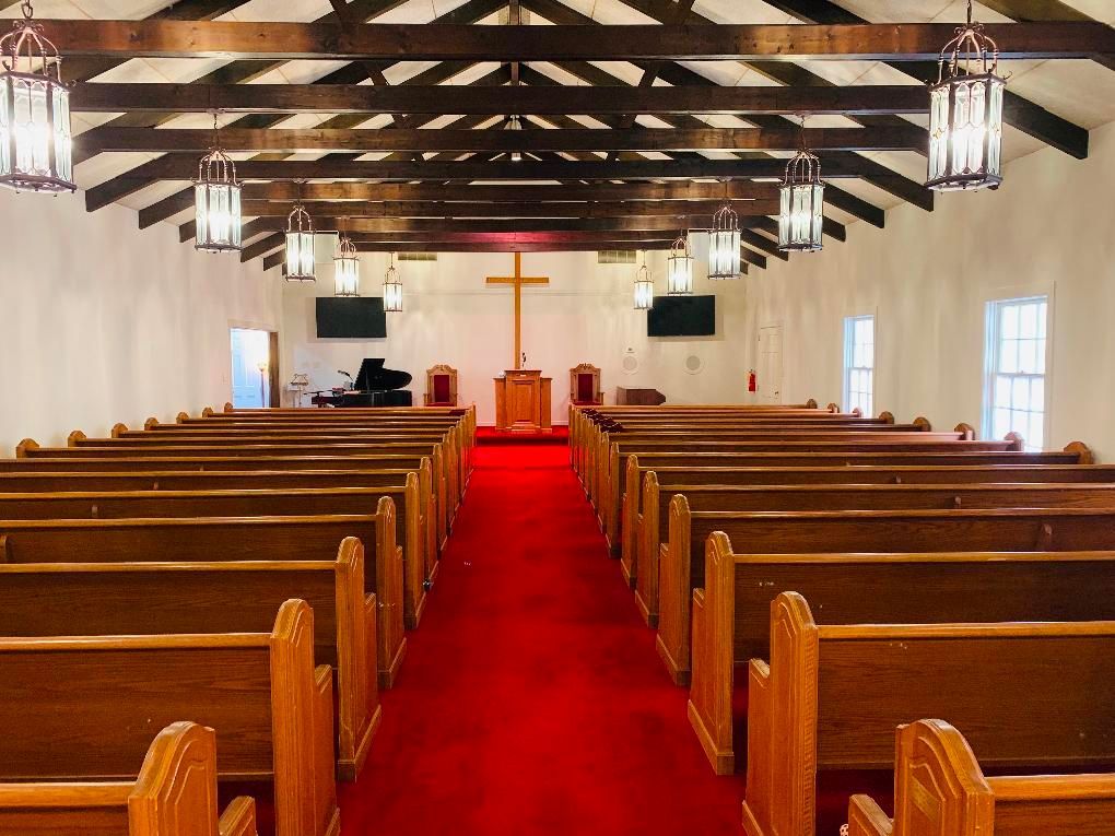 Interior of a small church with red carpet, wooden pews, and a cross at the front altar
