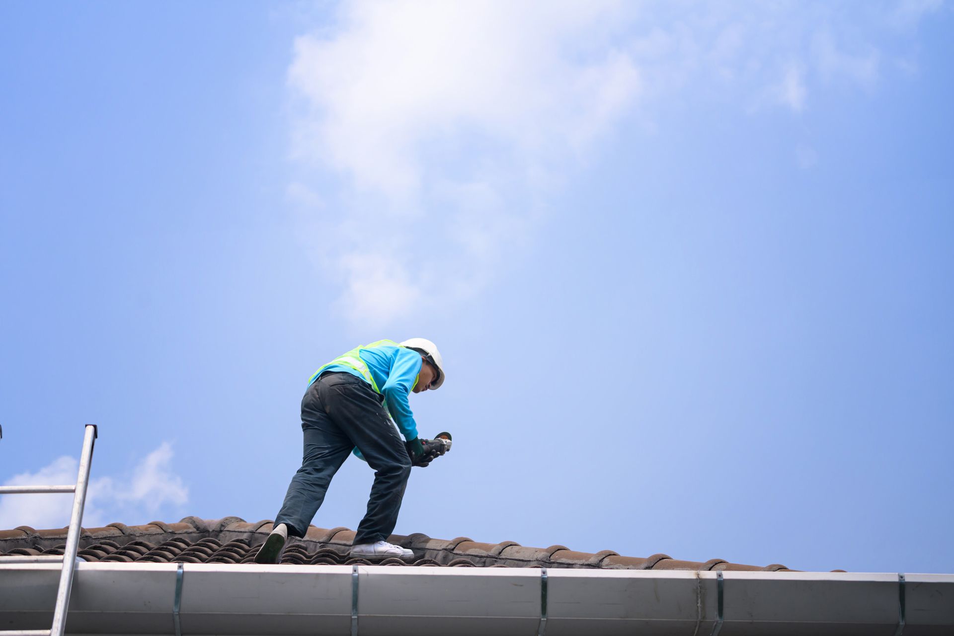 Roofer in safety gear installing tile roof on residential home against blue sky.