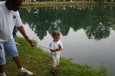 Man helping a child fish at a pond; green water, trees reflected, grassy bank.